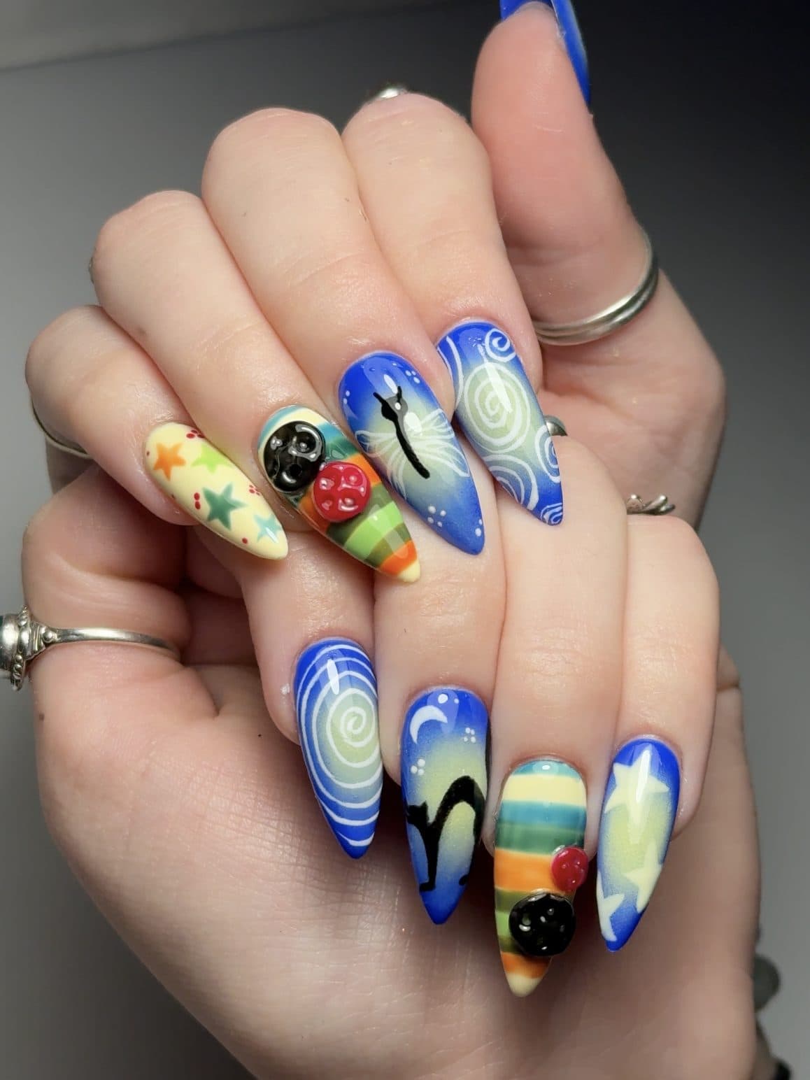 Bold maximalist almond Gel-X set with blue swirls, a black cat, fruit and 3D charms